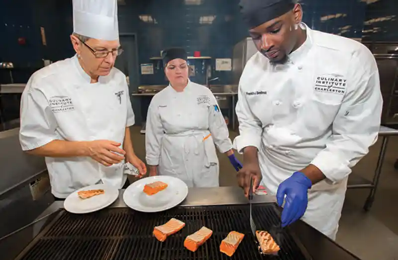 Two Culinary Institute of Charleston students cook salmon on the grill as an instructor looks on.